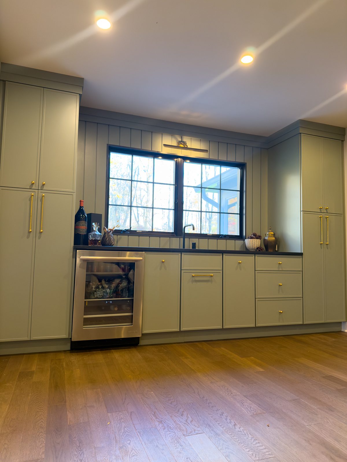 Sage-green bar with brass pulls, a black counter and an undercounter beverage fridge