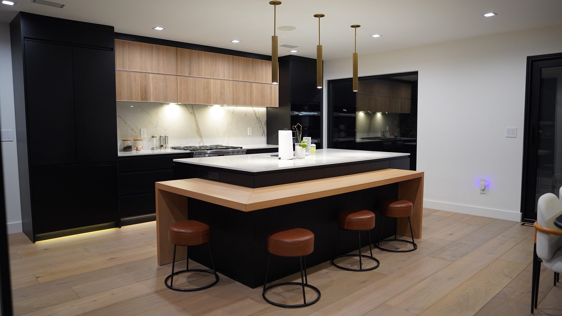 Matte black handleless kitchen with natural oak slab uppers and a marble-look waterfall island with an oak bar ledge and four leather stools