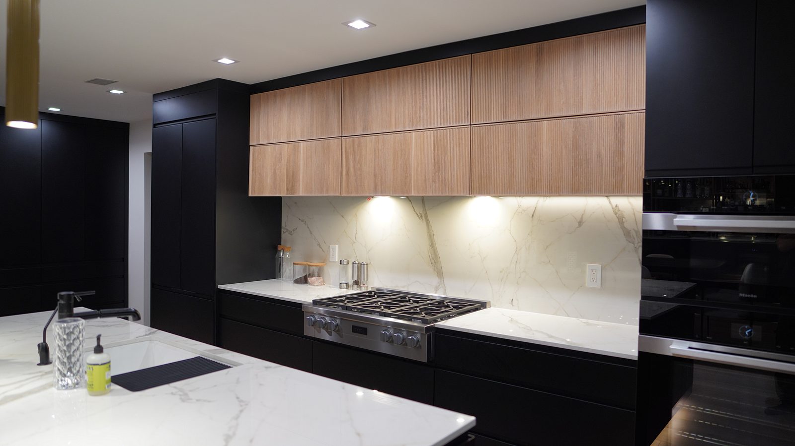 Cooktop wall: matte black base cabinets, oak uppers and the marble-look slab