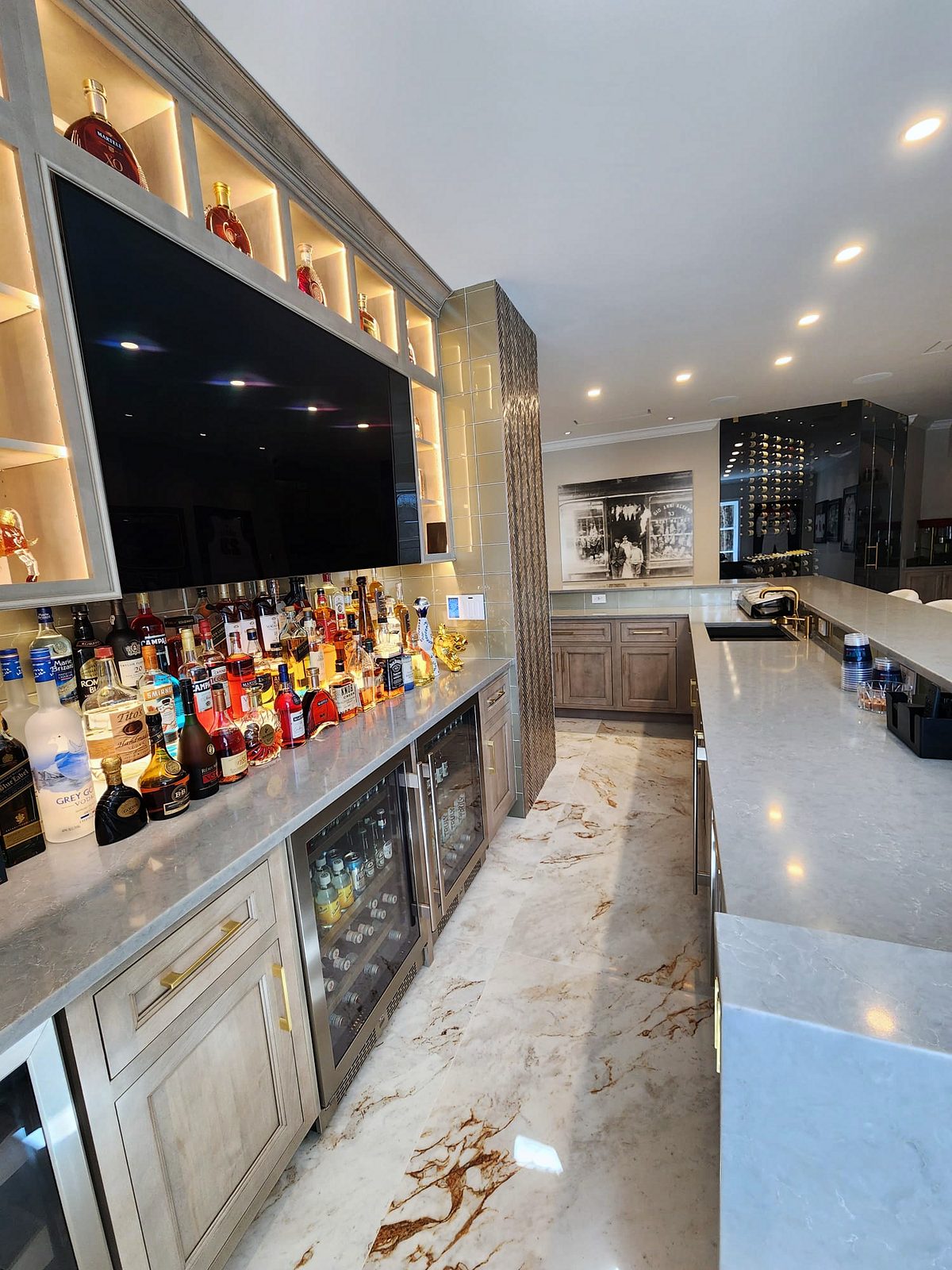 Bar wall with a television, bottle display and beverage fridges — Shiloh Aspen inset kitchen in a brown stain with a lit bar wall and a butler’s pantry