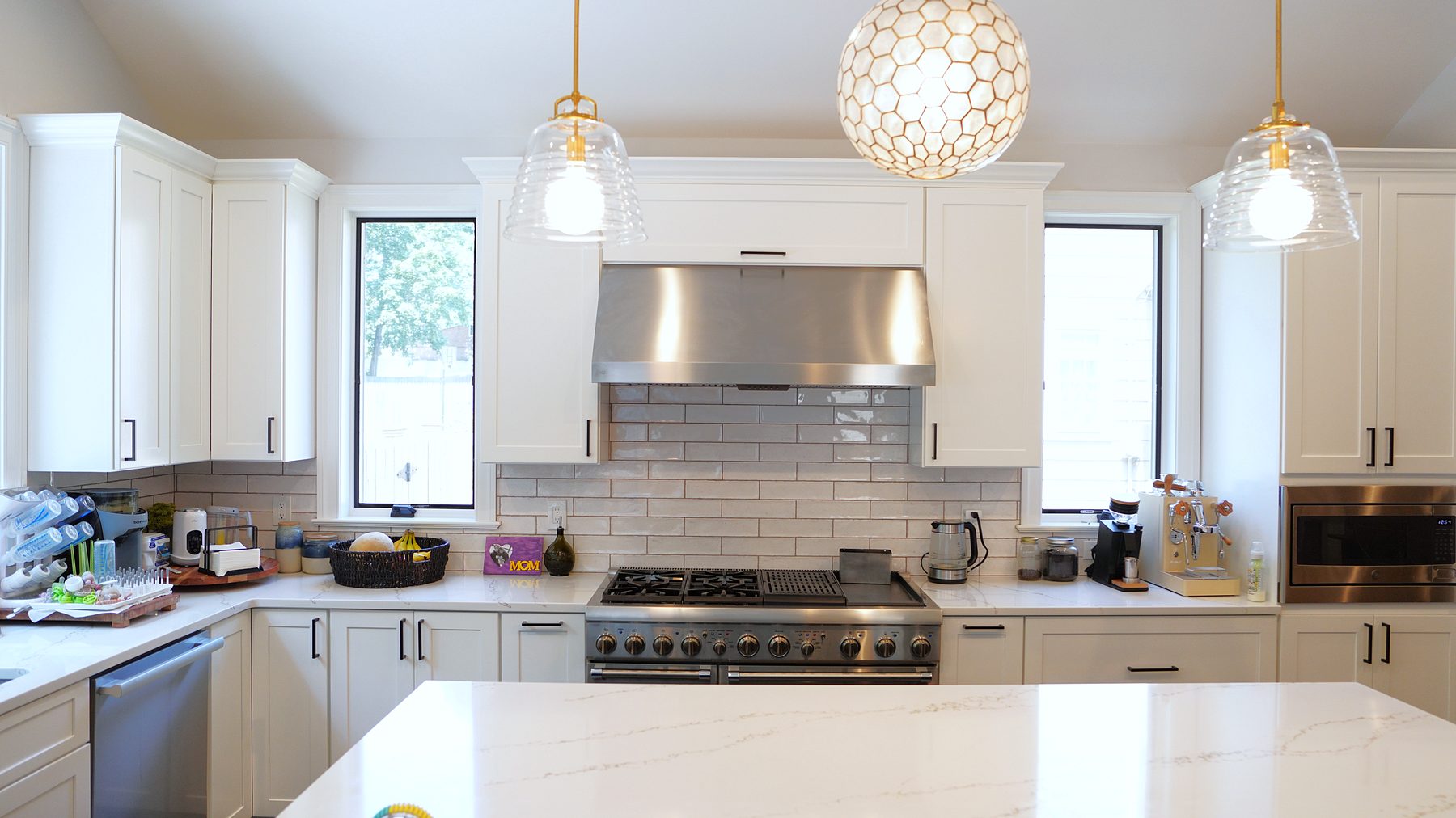 White shaker kitchen under a vaulted ceiling with a pro-style range, a stainless hood, black-framed windows and brass-trimmed glass pendants
