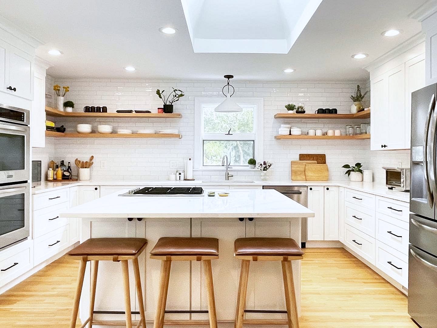 White island with a cooktop and leather saddle stools — White shaker kitchen with floating shelves and subway tile to the ceiling