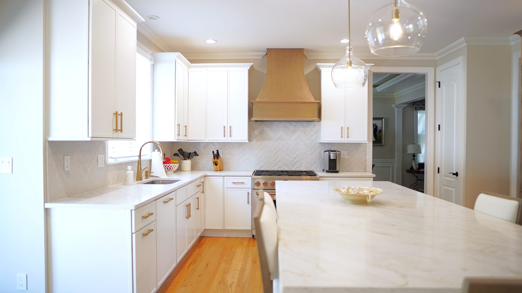 White shaker kitchen with a natural wood hood, a marble herringbone backsplash and a marble-look quartz island under glass globe pendants